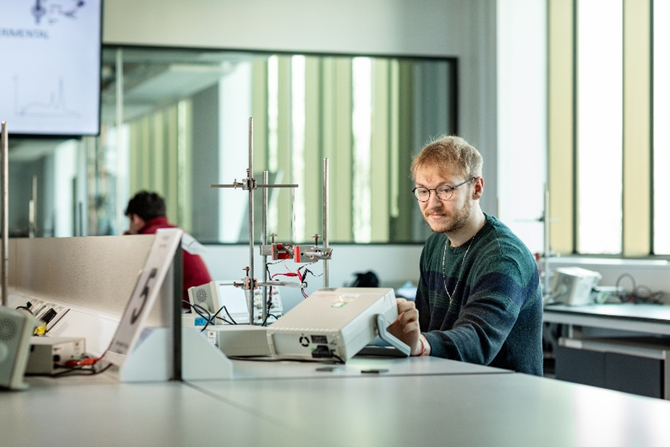 a male student sits at a lab bench looking at a piece of experimental equipment on the bench.